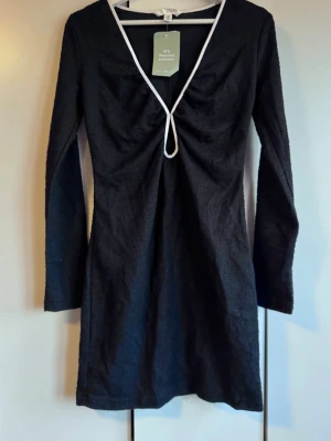 Ny Svart kort klänning. S. H&M. NWT. - Svart kort klänning från H&M med vit kant längs den djupa v-ringningen. Klänningen har lång ärm och är tillverkad av återvunnen polyester. Snygg detalj med rynkning framtill vid bysten. Oanvänd. Struktur i tyget typ boucle men tunnare och stretchigt. Dragkedja i ena sidan. Storlek S. Från H&M. Lappen kvar. Finns inget att anmärka på. NWT.