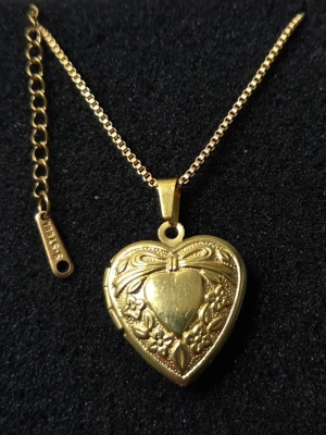 Guldfärgat hjärthalsband med medaljong Rostfritt Stål  - Snyggt guldfärgat halsband från Stiles med hjärtformad medaljong. Medaljongen har fina blommönster och en rosett på framsidan, och går att öppna för att lägga in ett litet foto eller minne. Kedjan är tunn och elegant med justerbar längd.                                             Detaljer: Material: Rostfritt stål (S-steel) Färg: Guldtonad Design: Hjärtformad medaljong med rosett och blomsterdekor Kedjans längd: 45 cm + 5 cm förlängningskedja Hänge: Ca 2 cm Skick: Nytt