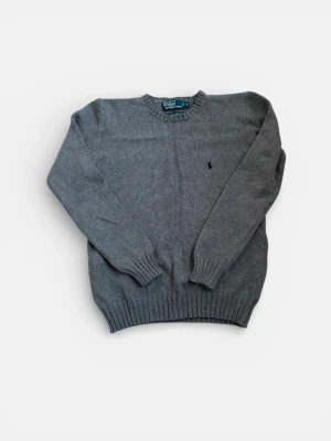 Grå stickad tröja från Polo Ralph Lauren - Klassisk grå stickad tröja från Polo Ralph Lauren med rund halsringning och ribbade muddar. Tröjan har den ikoniska mörkblå broderade loggan på bröstet och är tillverkad i mjukt material som passar perfekt till lager på lager.