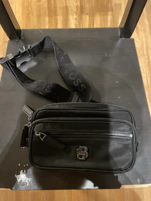 Svart axelväska från BOSS - Snygg svart axelväska från BOSS med två dragkedjefack och justerbar axelrem med BOSS-logga. Väskan har en stilren design med metallogo framtill och är tillverkad i slitstarkt material. Perfekt för att bära det viktigaste med stil.