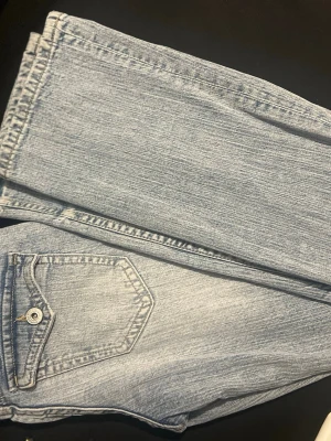 Ljusblåa jeans  - Säljer ett par ljusblå jeans med rak passform och unik ficka med lock och knapp framtill. Jeansen har klassisk denimlook och är tillverkade i bomull. Perfekta för dig som gillar en avslappnad stil helt nya aldrig använda 