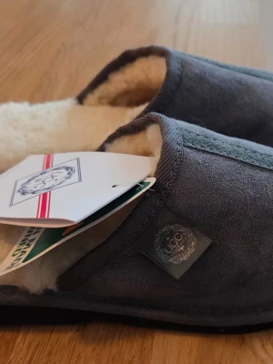 Grå fårskinnstofflor från UGG - Mysiga grå tofflor från UGG med utsida i mocka och insida i mjukt, vitt fårskinn. Klassisk slip-in modell med rund tå och diskret UGG-logga på sidan. Perfekta för att hålla fötterna varma hemma.