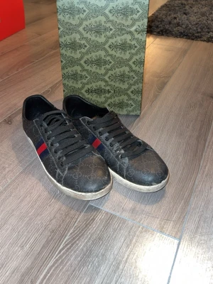 Svarta Gucci sneakers med röd/blå detalj - Snygga svarta sneakers från Gucci med klassiskt GG-mönster över hela skon och en röd/blå rand på sidan. Skorna har svarta skosnören och vit sula. Tillverkade i läder med textilinslag och låg profil som passar till streetwear.