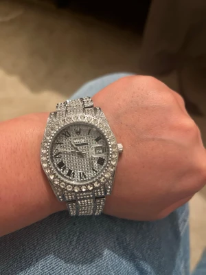 Iced out silverfärgad klocka - Säljer en riktigt blingad klocka från Wwoor i silverfärg med massor av glittrande stenar över hela boetten och armbandet. Klockan har romerska siffror, datumvisning och en chunky design som verkligen sticker ut.