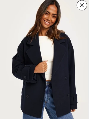 Blå kappa från NELLY - Snygg mörkblå kappa med dubbelknäppning och breda slag. Modellen är boxig och har stora knappar samt fickor på sidorna. Perfekt att slänga över en t-shirt och jeans för en cool look. Materialet ser ut att vara ullblandning.