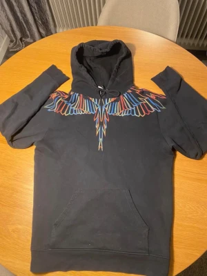 Svart hoodie med färgglada vingar - Säljer en svart hoodie från Marcelo Burlon med ikoniskt färgglatt vingmönster över bröstet och axlarna. Hoodien har huva med dragsko och en stor magficka. Perfekt för dig som vill sticka ut med en unik och trendig look.