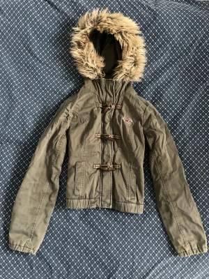 Vintage olivgrön parkajacka från Hollister - Snygg vintage olivgrön parkajacka från Hollister med fuskpäls på huvan och träknappar framtill. Jackan har långa ärmar, två fickor och en klassisk Hollister-logga på bröstet. Perfekt för kyliga dagar och riktigt trendig look. Vintage storlek L passar mer som s-m. Måtten finns. 