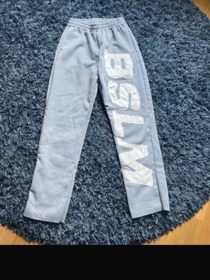 Grå mjukisbyxor med BSLM-tryck - Snygga grå mjukisbyxor med stort vitt BSLM-tryck längs ena benet och en cool stjärnliknande detalj på baksidan av benet. Byxorna har resår i midjan och är gjorda i mjukt sweatshirtmaterial, perfekta för chill dagar.