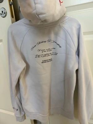 Vit hoodie från Hoodrich med tryck - Snygg vit hoodie från Hoodrich med stor svart logga och röd detalj på bröstet. Har huva med dragsko, magficka med dragkedja och texttryck på ryggen. Perfekt för dig som gillar streetwear och vill sticka ut lite extra.