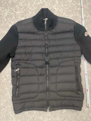 Moncler cardigan - Snygg svart cardigan från Moncler med quiltad front och stickade ärmar samt rygg. Jackan har hög krage, två dragkedjefickor och Moncler-logga på ena ärmen. Perfekt för kalla dagar och ger en clean, modern look. Den har blivit älthetsverifierad av vinted i deras egna betal tjänst. Den är i storlek M, killen på bilden är ungefär 180cm. Priset kan diskuteras, kan ta byten.