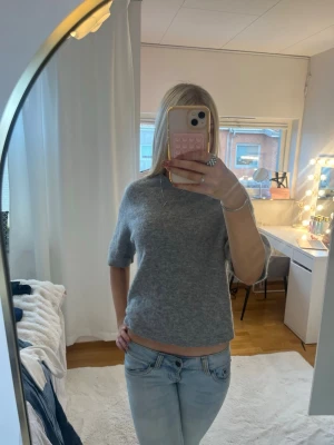 Grå stickad tröja med kort ärm - Mysig grå stickad tröja med korta ärmar och rund hals. Tröjan har en enkel och stilren design som passar perfekt till jeans eller kjol. Materialet ser ut att vara mjukt och bekvämt, perfekt för lager på lager under kyligare dagar.