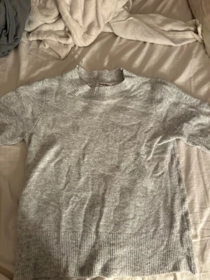 Ljusgrå stickad tröja från J'aime - Mysig ljusgrå stickad tröja från J'aime med rund halsringning och breda ribbade muddar vid ärmslut och nederkant. Tröjan har en klassisk rak passform och är perfekt för lager-på-lager-stil under kyliga dagar. Köpt från missäy