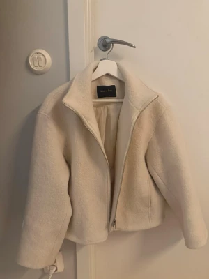 Beige ullcardigan från Massimo Dutti - Stilren beige cardiganjacka från Massimo Dutti i mjuk ullmix. Jackan har dragkedja framtill, hög krage och raka ärmar. Perfekt för lager på lager och enkel att matcha med olika outfits.