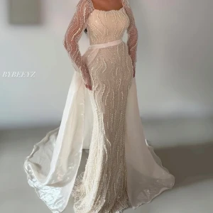 Lång off white / beige  klänning med pärlor - Elegant långklänning i vit nyans med stenar,pärldetaljer och transparenta långärmar. Klänningen har markerad midja med band och ett långt släp baktill. Perfekt för dig som vill sticka ut på bal eller fest.