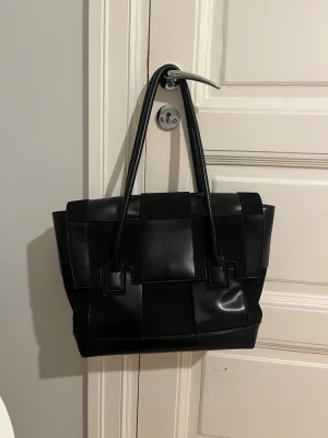 Svart flätad handväska  - Stilren svart handväska med flätad design och långa handtag. Perfekt för dig som gillar minimalistisk och trendig look. Finns små spår av användning