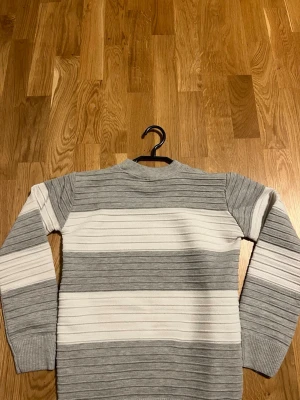 Gråvit randig stickad tröja - Säljer en stickad tröja med breda horisontella ränder i grått och vitt. Tröjan har rund halsringning, långa ärmar och ribbstickade muddar. Materialet är mjukt och stretchigt, perfekt för kyligare dagar.