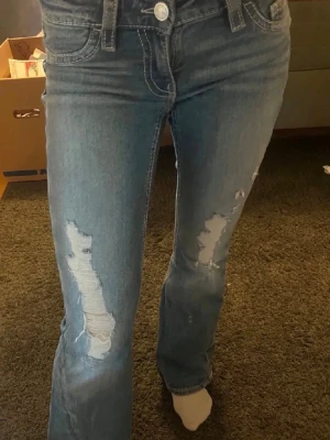 Blå slitna bootcut jeans - Säljer ett par ljusblå bootcut jeans med slitningar och hål på både lår och knän. Jeansen har låg midja, klassiska fem fickor och vita kontrastsömmar. Ifrån Gina Tricot i storlek xs 