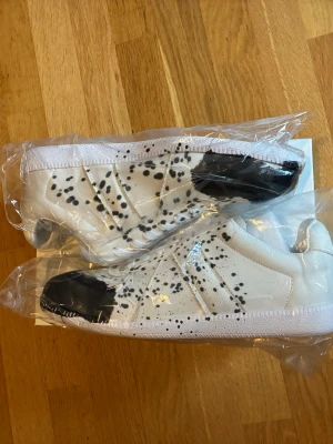 Masion margiela. Vita sneakers  - Unika vita sneakers med svart stänkmönster och svarta detaljer på tå och häl. Skorna har en klassisk låg siluett och är tillverkade i textil med gummisula. Perfekta för dig som vill sticka ut med en kreativ och trendig look.