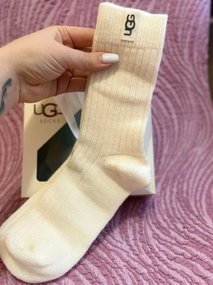 Vita ribbade strumpor från UGG - Säljer ett par vita ribbade strumpor från UGG. Strumporna är höga, har en mjuk och stretchig känsla och är perfekta för att hålla fötterna varma. Klassisk UGG-logga upptill och stilren design.