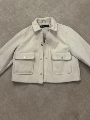 Beige kappa från Zara - Stilren beige kappa  från Zara med två stora bröstfickor och tryckknappar framtill. Kappan har en rak, boxig passform och klassisk krage. Materialet är mjukt och känns ulligt, perfekt för lager på lager. Enkel att matcha till olika outfits. Perfekt för våren och hösten! Dock så har den vänstra knappen på fickan gått av så det går inte att stänga den helt men den funkar bra annars! 