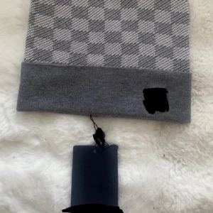 Gråvit mönstrad mössa med uppvikt kant - Snygg grå och vit mössa med diskret rutmönster och bred uppvikt kant. Mössan har en stilren look och passar perfekt till kalla dagar. Märke syns på kanten. Materialet ser ut att vara mjukt och stretchigt, troligen bomull eller akryl.