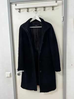 Etage kappa - Svart kappa från Etage med en strukturerad yta. Kappan har en klassisk krage och knäppning framtill. Den är designad med två diskreta fickor i sidorna. En mångsidig kappa som passar för olika tillfällen.