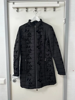 MICHA jacka - Svart jacka med ett detaljerat mönster. Jackan har en dragkedja framtill och två fickor med dragkedja. Insidan av jackan har ett färgglatt blommigt mönster med fåglar och fjärilar.