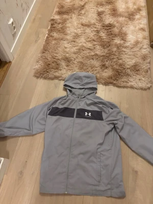 Grå vindjacka från Under Armour - Snygg grå vindjacka från Under Armour med svart panel och logga på bröstet. Jackan har huva med dragsko, hel dragkedja framtill och två sidofickor. Tillverkad i lätt och ventilerande material med meshfoder för extra komfort.
