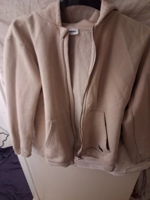 Beige hoodie från KappAhl med dragkedja - Beige hoodie från KappAhl med dragkedja framtill och två stora fickor. Insidan är fodrad med mjukt teddyfoder för extra värme och komfort. Klassisk huva och ribbade muddar vid ärmslut och nederkant. Perfekt för en avslappnad och stilren look.