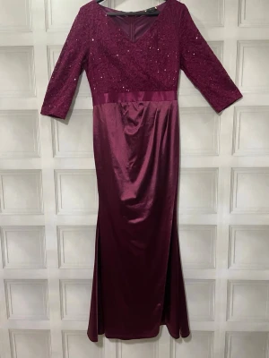 Vinröd långklänning med spets och paljetter - Elegant vinröd långklänning med v-ringning, spets och paljetter upptill samt trekvartsärm. Kjolen är i glansigt tyg med slits på sidorna och markerad midja med satinband. Perfekt för speciella tillfällen.