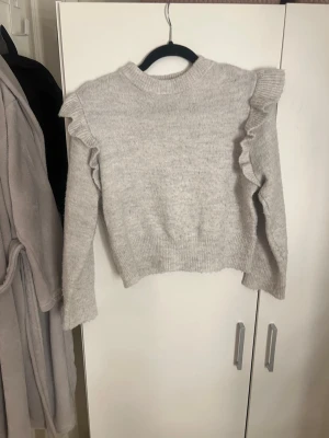 Ljusgrå stickad tröja med volang - Supersöt ljusgrå stickad tröja från Gina Tricot med volangdetaljer längs axlarna. Tröjan har ribbade muddar vid ärmslut och nederkant samt en högre rund hals. Perfekt för dig som gillar en trendig och mysig look.