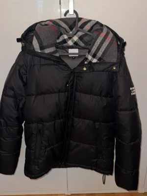 Svart pufferjacka från Burberry - Säljer en svart pufferjacka från Burberry med klassiskt rutigt foder i grått, vitt och rött. Jackan har huva, dragkedja framtill, två fickor med dragkedja och logga på ärmen. Perfekt för kalla vinterdagar och riktigt snygg streetstil.