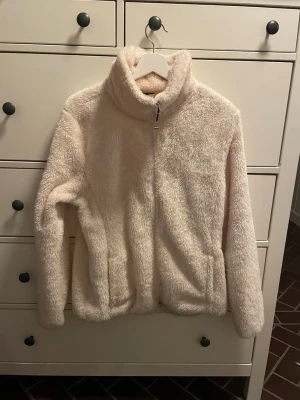 Beige teddyjacka  - Mysig beige teddyjacka med hög krage och dragkedja framtill. Jackan har två fickor på sidorna och är gjord i fluffigt, mjukt material som håller dig varm. Perfekt för kyliga dagar och ger en avslappnad, trendig look. Köpt på hööks. Märke CRW.