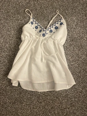 Vit broderad linnetopp med knyt - Söt vit linnetopp med tunna axelband och blå broderade blommor vid urringningen.  Perfekt för varma dagar och ger en fräsch sommarkänsla. Det finns ingen lapp så märke eller storlek vet jag ej, men skulle gissa på xs-s