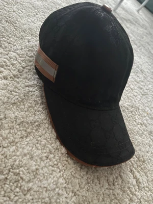 Svart GC keps med beige detalj - Säljer en svart keps från GC med diskret GC-mönster i tyget och en beige och grå rand på sidan. Kepsen har böjd skärm och är tillverkad i bomull. Märket syns tydligt på insidan och den har en snygg, klassisk look. Säljer så billigt för behöver pengar