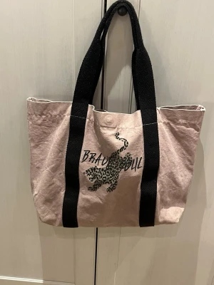 Lila shoppingväska med leopardtryck - Snygg rosa shoppingväska i tyg med svarta breda handtag. På framsidan finns ett leopardmönstrat tryck och texten 'BRAVO DUL'. Väskan har en rymlig insida med innerficka för småsaker. Perfekt för dig som gillar trendiga accessoarer.
