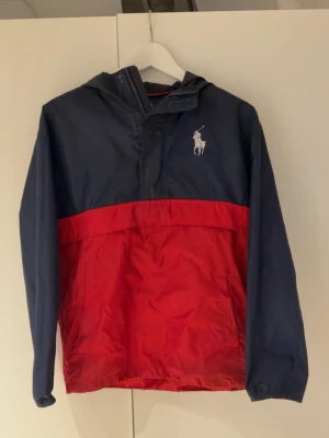 Polo Ralph Lauren vindjacka marin/röd - Snygg vindjacka från Polo Ralph Lauren i marinblått och rött. Jackan har huva, stor logga på ryggen och klassisk Polo-häst på bröstet. Tillverkad i lätt och tåligt material, perfekt för blåsiga dagar. Sportig och trendig look.