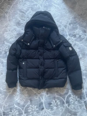 Svart pufferjacka från Moncler - Snygg svart pufferjacka från Moncler med stor huva och två dragkedjefickor framtill. Jackan har en patch med Moncler-logga på ena ärmen och quiltad design. Perfekt för kalla dagar och riktigt trendig look.