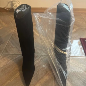 Svarta knähöga stövlar med klack - Säljer ett par svarta knähöga stövlar med spetsig tå och smal klack. Stövlarna har en stilren och elegant design i mockaliknande material. Perfekta för att lyfta vilken outfit som helst.