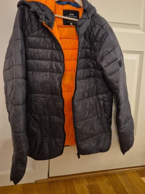 Grå dunjacka från Jack & Jones - Snygg grå dunjacka från Jack & Jones med orange foder och huva. Jackan har quiltad design, dragkedja framtill och är lätt vadderad för extra värme. Perfekt för kalla vinterdagar och ger en cool kontrast med det orangea innertyget.