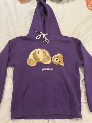 Lila hoodie från Palm Angels - Säljer en lila hoodie från Palm Angels med en stor broderad nallebjörn på bröstet och vit logga under. Hoodien har huva med vita dragsnören och en klassisk magficka. Materialet är mjukt och skönt, perfekt för en avslappnad streetwear-look.