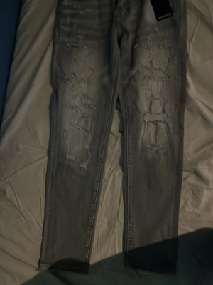 Grå slitna skinny jeans - Säljer ett par grå skinny jeans med slitningar och hål på framsidan av benen. Jeansen har en tight passform och klassisk femficksdesign. Perfekta för dig som gillar en edgy och trendig stil. 