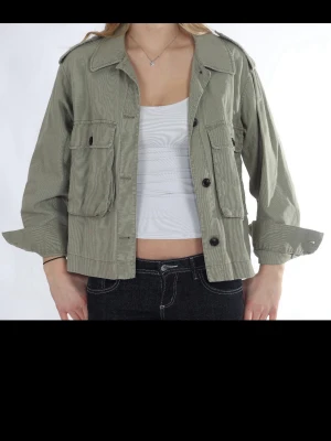 Grön utilityjacka med fickor - Snygg grön utilityjacka med stora bröstfickor och knappar framtill. Jackan har krage, långa ärmar och en loose passform som ger en avslappnad look. Perfekt att slänga över en t-shirt för en cool vardagsstil.