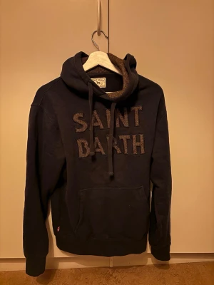 Mörkblå Saint Barth hoodie med huva - Säljer en svart hoodie från Saint Barth Country Club med stor broderad text 'SAINT BARTH' på bröstet. Hoodien har en stor huva med brunt fiskbensmönstrat foder och dragsnören samt en klassisk magficka framtill.