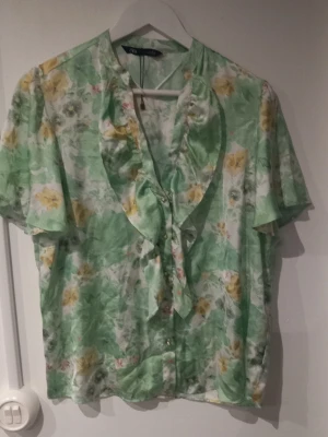 Grön blommig blus från Zara - Söt blus från Zara i ljusgrönt med blommigt mönster i gult och vitt. Blusen har korta ärmar, volangkant längs knäppningen och en luftig, lätt materialkänsla. Perfekt för dig som gillar romantiska och somriga vibes.