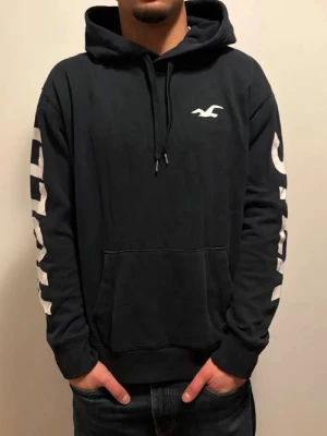 Sällsynt Svart hoodie från Hollister - Svart hoodie från Hollister med vit logga på bröstet och stora vita tryck på båda ärmarna. Klassisk känguruficka framtill och justerbar huva med snören. Tillverkad i mjukt bomullsmaterial som är skönt att ha på sig. EXTREMT EFTERTRAKTAD 