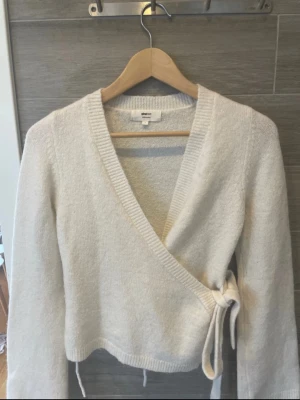 Vit omlott kofta med knyt - Säljer en supermjuk vit omlott kofta med knyt i sidan och v-ringad hals. Koftan har långa ärmar och är stickad i ett fluffigt material som känns riktigt cozy. Perfekt att slänga på sig när det är lite kyligt ute.