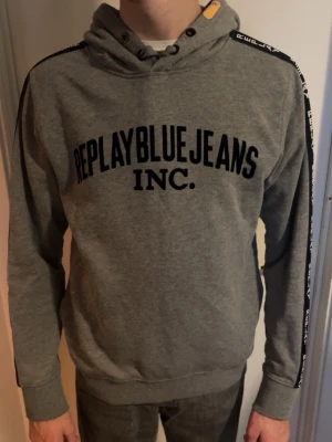Grå hoodie från Replay Blue Jeans - Snygg grå hoodie från Replay Blue Jeans med svart tryck framtill och svarta band med Replay-logga längs ärmarna. Strlk M. Gott skick. Pris kan diskuteras.