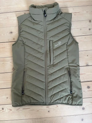 Olivgrön dunväst från Pelle P - Snygg olivgrön dunväst från Pelle P med dragkedja framtill, hög krage och tre praktiska fickor med dragkedja. Västen har ett quiltat mönster och är tillverkad i ett tåligt material som håller värmen. Perfekt för lager på lager och outdoor-stil.                                                        Riktigt bra kvalitet och är inte använd.