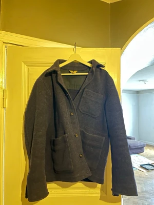 Mörkblå ullrock från Menchi Tessuti - Stilren mörkblå ullrock från arket Menchi Tessuti serie med tre stora utanpåfickor och klassisk krage. Rocken har knappstängning framtill och är helfodrad med svart innerfoder. Perfekt för dig som gillar tidlös och enkel design.